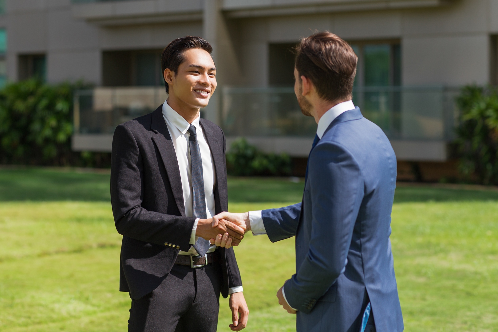 Closeup of two smiling business men shaking hands and standing on lawn with building in background